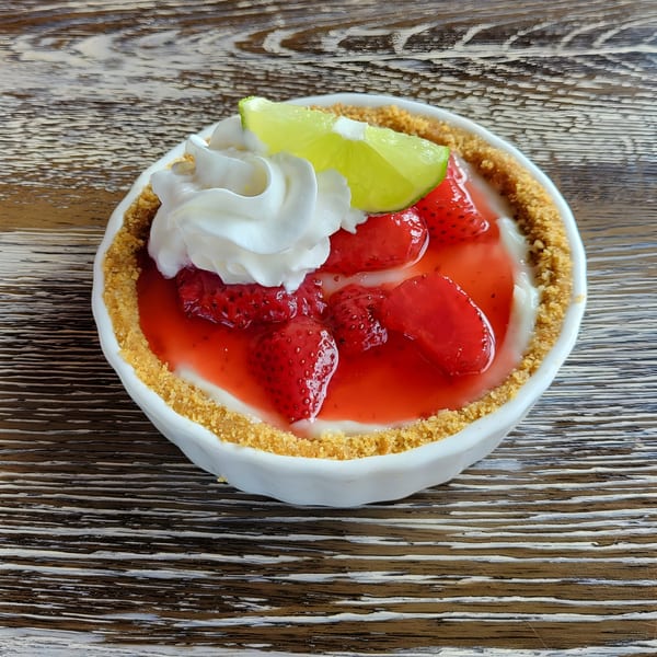 Strawberry cheesecake with whipped cream and a lime wedge in a white bowl on a wooden table.