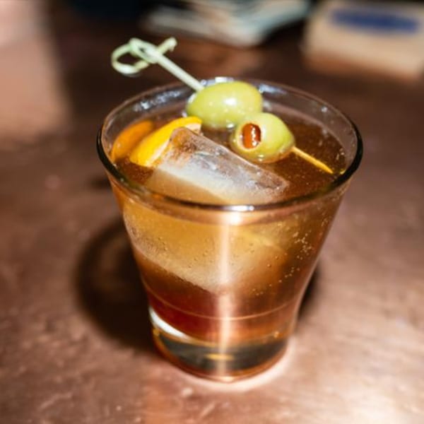 A Latin fusion cocktail with olives and an orange slice, served in a rocks glass.