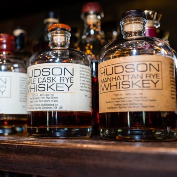 Close-up of two bottles of Hudson whiskey, Manhattan Rye and Maple Cask Rye, on a dark shelf.