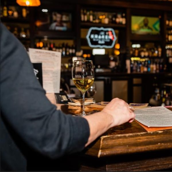 A person's arm and hand resting on a wooden bar next to a glass of white wine.