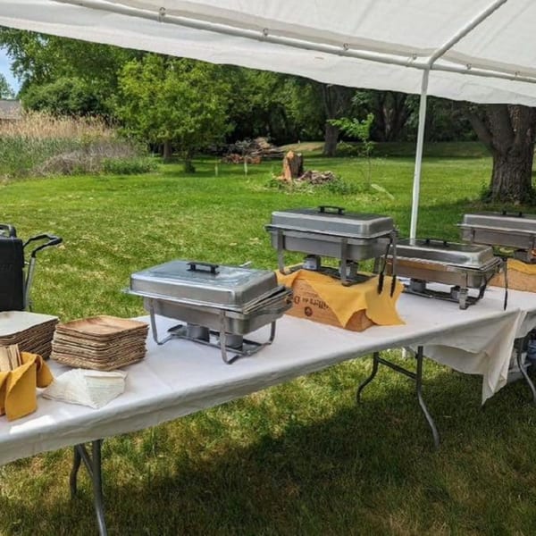 Outdoor catering setup with chafing dishes, plates, and napkins under a white tent on green grass.