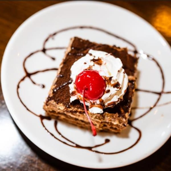 A square chocolate dessert with whipped cream and a cherry on a white plate with chocolate drizzle.